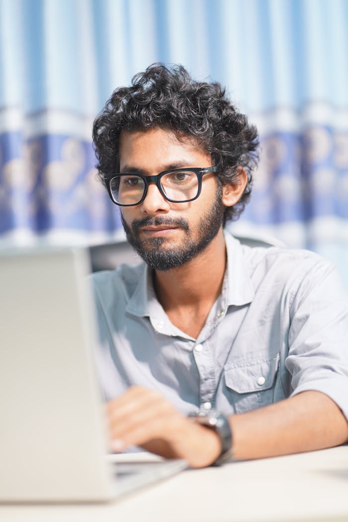 A South Asian adult male deeply focused on working on his laptop indoors.