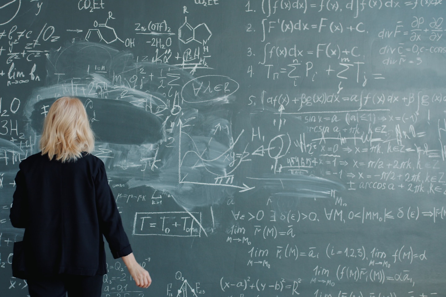 Back view of female scientist walking to chalkboard writing formulas with chalk concentrated on science. Occupation, intelligent people and knowledge concept.